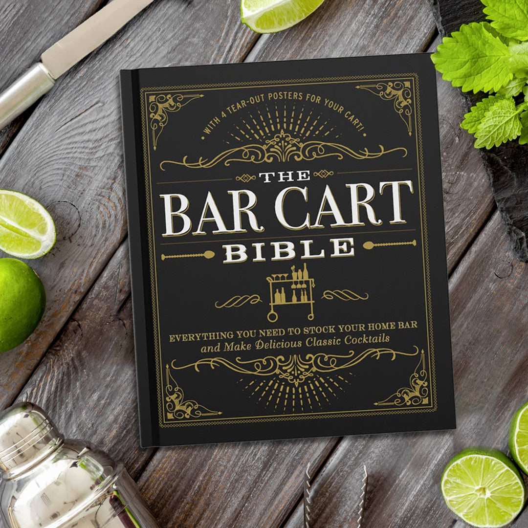 Book titled 'The Bar Cart Bible' on a wooden surface with limes and a cocktail shaker.