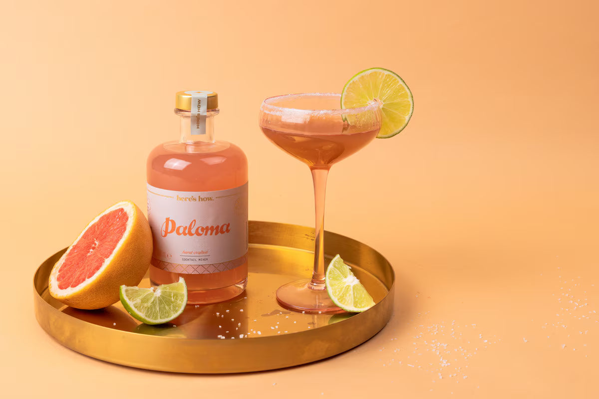 Bottle of Paloma cocktail with a glass and ingredients on a gold tray against an orange background