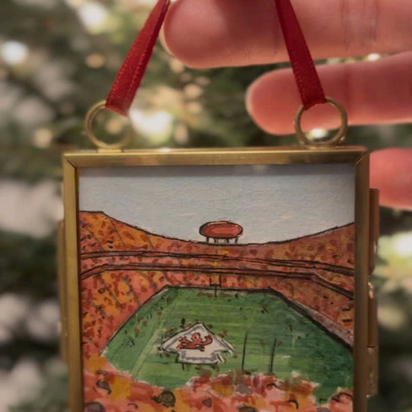 A hand holds a square gold-toned pendant necklace featuring a hand-painted miniature scene of a football stadium in autumn colors. The pendant is shown against a blurred background of green foliage.