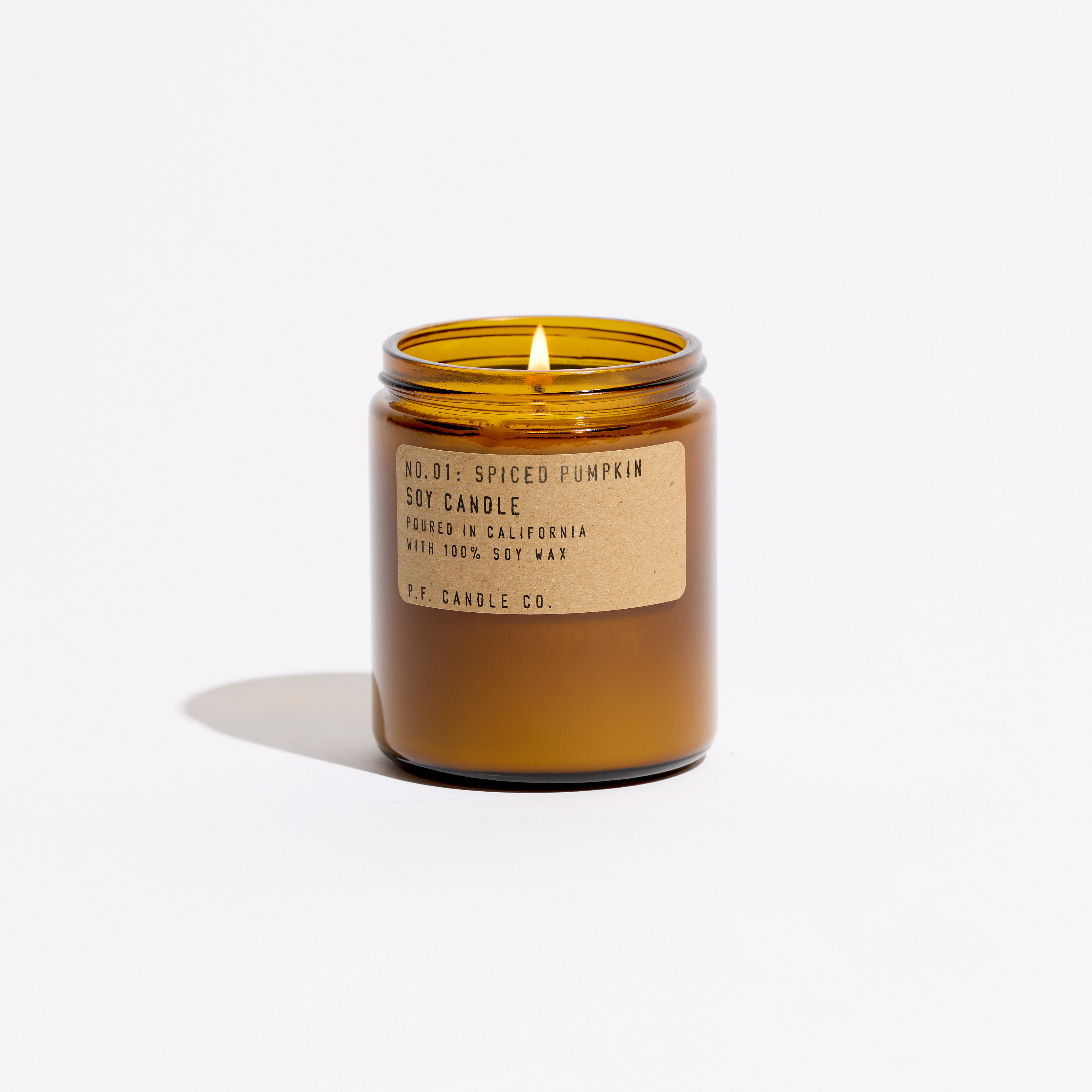 An amber glass jar candle with a lit wick, featuring a beige kraft paper label that reads 'No. 01: Spiced Pumpkin Soy Candle' and 'P.