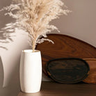 White vase with dried pampas grass against a neutral wall with wooden elements.