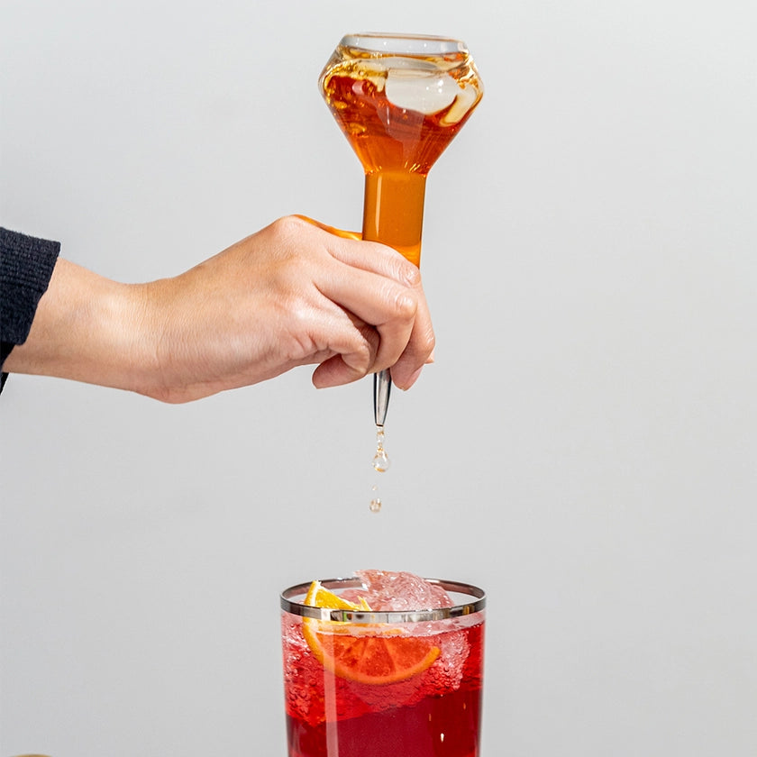 Hand using a honey dripper to add honey to a glass of red liquid with ice and orange slices.