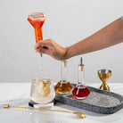 Hand pouring a red liquid from a bottle into a glass on a marble tray with other bottles and a cocktail.