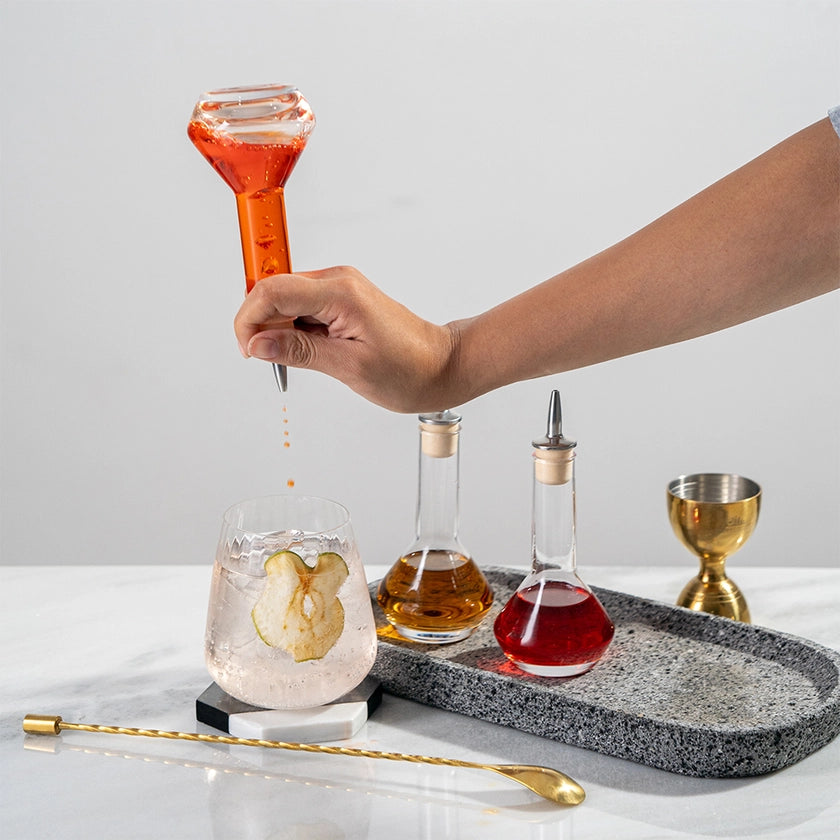 Hand pouring a red liquid from a bottle into a glass on a marble tray with other bottles and a cocktail.