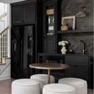 A modern interior scene showing a black refrigerator, black cabinetry, a round wooden table, three white textured poufs, and a staircase with white steps and a light wood handrail against a white wall.