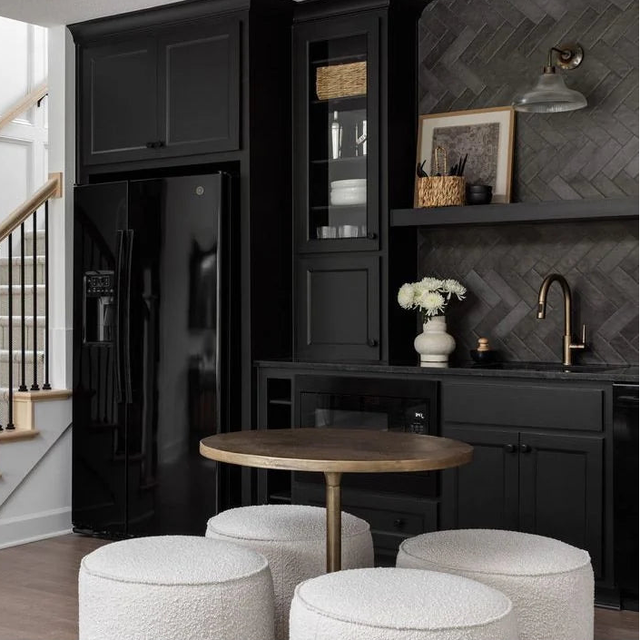 A modern interior scene showing a black refrigerator, black cabinetry, a round wooden table, three white textured poufs, and a staircase with white steps and a light wood handrail against a white wall.