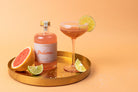 Bottle of Paloma cocktail with a glass and ingredients on a gold tray against an orange background