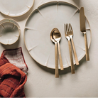 Table setting with ceramic plates, gold cutlery, and a small cup on a textured surface.