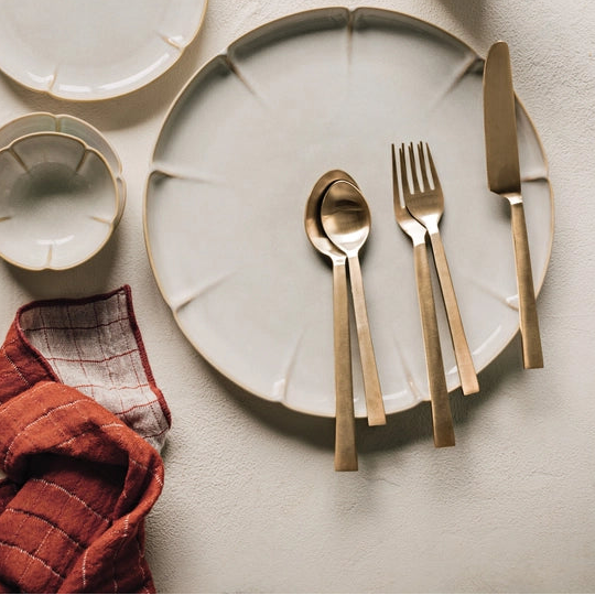 Table setting with ceramic plates, gold cutlery, and a small cup on a textured surface.