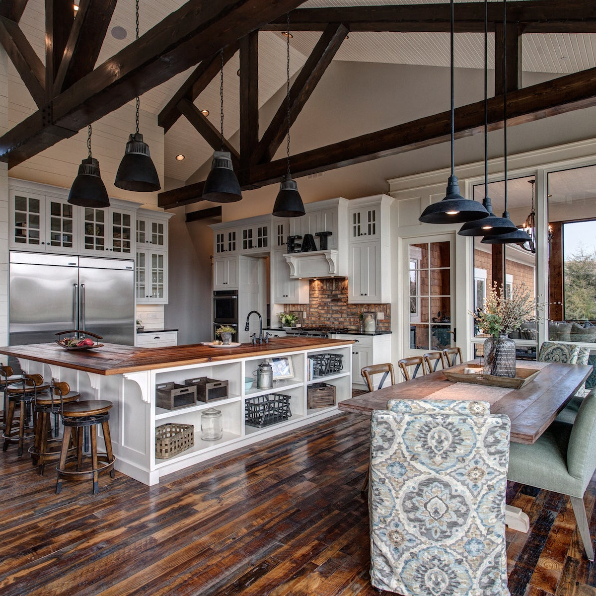 A high-ceilinged modern kitchen with dark wood ceiling beams, white cabinetry, a central island with wood countertops, pendant lights, and a dining area with patterned chairs and a wooden table.