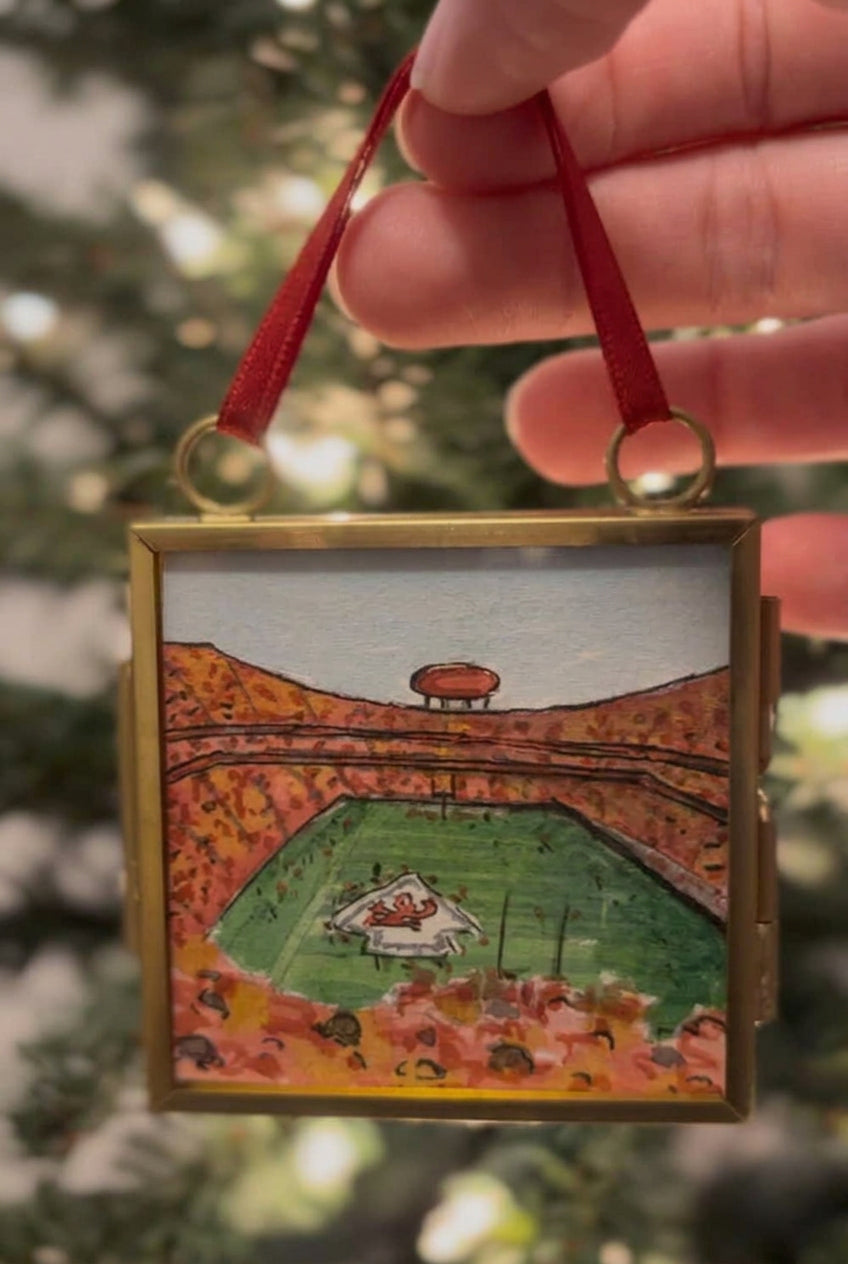 A hand holds a square gold-toned pendant necklace featuring a hand-painted miniature scene of a football stadium in autumn colors. The pendant is shown against a blurred background of green foliage.