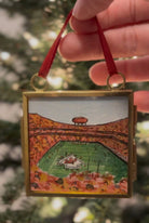 A hand holds a square gold-toned pendant necklace featuring a hand-painted miniature scene of a football stadium in autumn colors. The pendant is shown against a blurred background of green foliage.