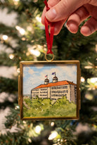 A hand holds a square gold-tone pendant necklace featuring a watercolor painting of a historic building with a red-orange roof and green foreground, suspended from a gold chain against a blurred outdoor background.