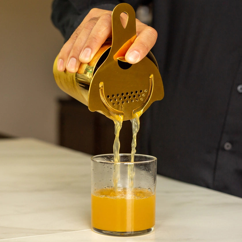 Gold manual juicer squeezing orange juice into a glass on a neutral background