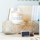 Candle with 'furnish' branding on a table with books and a basket.